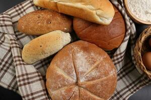 frisch gebacken Brot Laib Brötchen rollen runden lange mischen Wahrheit eingewickelt im kariert Küche Stoff Serviette Handtuch Weizen Blume Öl Wasser Salz- Eier rollen Stift Schiefer Stein Über schwarz Hintergrund foto