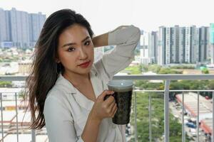 schön jung Süd-Ost asiatisch Frau halten Tee Kaffee Tasse in der Nähe von Balkon Fenster Stadt scape Hintergrund, suchen entspannend Denken foto