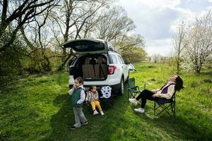 Mutter mit zwei Töchter sitzen auf Stuhl gegen Auto öffnen Kofferraum auf Picknick. foto