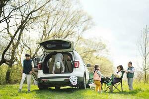 Mutter mit Kinder sitzen auf Stuhl gegen Auto öffnen Kofferraum auf Picknick. foto