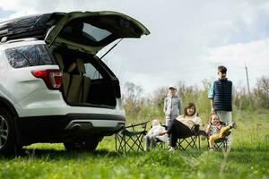 Mutter mit Kinder sitzen auf Stuhl gegen Auto öffnen Kofferraum auf Picknick. foto