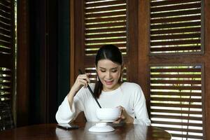 jung asiatisch Frau sitzen beim rustikal Cafe Essen von Chinesisch Essstäbchen von Suppe Nudel von Schüssel aussehen Nieder beim Essen foto