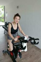 schön attraktiv Mitte Alter Süd-Ost asiatisch Frau ausüben stationär Fahrrad Zyklus Fahrrad im ein Gymnasium gesund Lebensstil aussehen nach vorne Lächeln beim Kamera foto