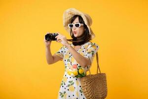 Studio Porträt jung ziemlich asiatisch Frau glücklich Lächeln Dressing Frühling Mode mit Sonnenbrille, gewebte Tasche und halten Tulpe Strauß Blume und Kamera gegen Gelb Studio Hintergrund. Reise Konzept foto