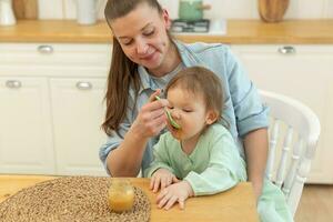 glücklich Familie beim heim. Mutter Fütterung ihr Baby Mädchen von Löffel im Küche. wenig Kleinkind Kind mit unordentlich komisch Gesicht isst gesund Essen beim heim. jung Frau Mama geben Essen zu Kind Tochter. foto