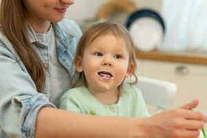 glücklich Familie beim heim. Mutter Fütterung ihr Baby Mädchen von Löffel im Küche. wenig Kleinkind Kind mit unordentlich komisch Gesicht isst gesund Essen beim heim. jung Frau Mama geben Essen zu Kind Tochter. foto