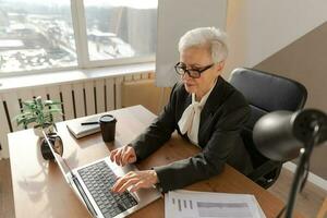 zuversichtlich stilvoll europäisch Mitte alt Senior Frau mit Laptop beim Arbeitsplatz. stilvoll älter reifen 60er Jahre grau behaart Dame Geschäftsfrau Sitzung beim Büro Tisch. Boss Führer Lehrer Fachmann Arbeiter. foto