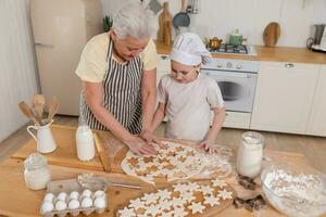 glücklich Familie im Küche. Oma Enkelin Kind Schneiden Kekse von Teig auf Küche Tabelle zusammen. Oma Lehren Kind Mädchen Koch backen Kekse. Haushalt Zusammenarbeit Portion Familie Generationen. foto