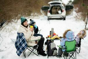 jung Frau mit Kinder im Winter Wald auf ein Picknick. Mutter und drei Kinder gegen Sie Auto mit öffnen Stamm. foto