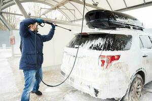 Mann wäscht Hochdruckwasser amerikanisches SUV-Auto mit Dachgepäckträger bei Selbstbedienungswäsche bei kaltem Wetter. foto