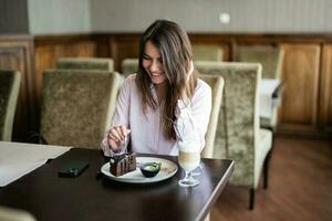 jung lächelnd Brünette Frau sitzen im Kaffee Geschäft Cafe Restaurant drinnen und Essen Schokolade Brownie Dessert Kuchen. foto