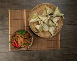 traditionell trocken Rindfleisch Curry Rendang im Kokosnuss Schale mit Diamant geformt gewebte Palme Blatt Beutel gedämpft glutenhaltig Reis Kuchen Ketupat kupat tipat auf Bambus Tablett Matte rustikal hölzern Tabelle Hintergrund foto