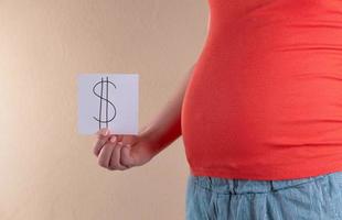 eine Nahaufnahme des Bauches einer schwangeren Frau in Rot, die ein weißes Blatt Papier mit einem Dollarsymbol hält
