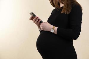junge schwangere Frau in einem schwarzen Kleid hält ihren Bauch und schaut auf ihr Handy foto