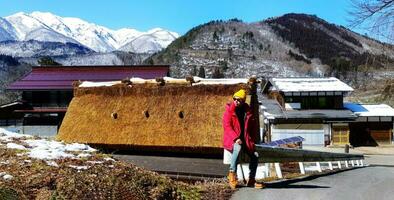 asiatisch Tourist Mann im rot Mantel tragen Sonnenbrille und Gelb stricken Hut Sitzung auf Stahl Schiene mit uralt Zuhause oder Haus, Berg und Himmel Hintergrund im japanisch Dorf, Japan. Asien reisen, Tourismus foto