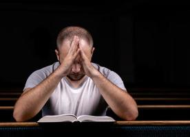Ein junger Mann sitzt auf einer Kirchenbank, liest die Bibel und betet