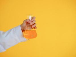 Wissenschaftler im weißen Kleid, der einen Becher der orange chemischen Lösung auf dem gelben Hintergrund hält foto