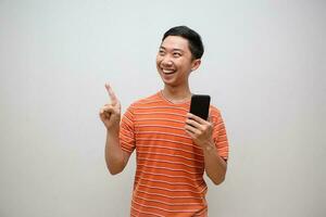 heiter asiatisch Mann Orange Hemd fühlt sich glücklich halten Handy, Mobiltelefon Telefon und Geste Punkt Finger isoliert foto