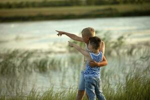 zwei Jungs im Jeans und T-Shirts Punkt ein Finger in das Entfernung in der Nähe von das Teich. Sommer- sonnig Tag in der Nähe von das Teich. Sommer- Ferien im das Landschaft. glücklich Kindheit foto