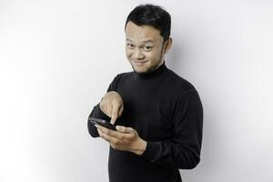 aufgeregt asiatisch Mann tragen schwarz Hemd lächelnd während halten seine Telefon, isoliert durch Weiß Hintergrund foto