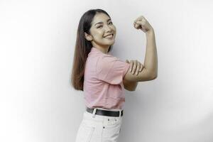 aufgeregt asiatisch Frau tragen ein Rosa T-Shirt zeigen stark Geste durch Heben ihr Waffen und Muskeln lächelnd stolz foto
