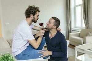 lgbtq Paar Umarmen jeder andere und schrumpft Wein drinnen. zwei romantisch jung männlich Liebhaber suchen beim jeder andere während Sitzung zusammen im ihr Leben Zimmer. jung Fröhlich Paar Sein romantisch beim heim. foto