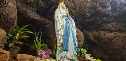 Statue von heilig Jungfrau Maria im römisch katholisch Kirche, im das Höhle von Jungfrau Maria, im ein Felsen Höhle Kapelle katholisch Kirche mit tropisch Blumen um foto