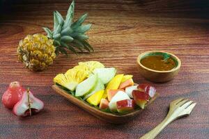 geschnitten Obst Salat, im Indonesien bekannt wie Rujak buah oder lutis oder Lotis. serviert mit würzig braun Zucker Soße und Boden Erdnüsse im ein Teller auf Holz Hintergrund foto