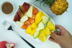 geschnitten Obst Salat, im Indonesien bekannt wie Rujak buah oder lutis oder Lotis. serviert mit würzig braun Zucker Soße und Boden Erdnüsse im ein Teller auf Weiß Hintergrund mit Hand foto