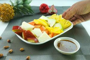 geschnitten Obst Salat, im Indonesien bekannt wie Rujak buah oder lutis oder Lotis. serviert mit würzig braun Zucker Soße und Boden Erdnüsse im ein Teller auf Weiß Hintergrund mit Hand foto