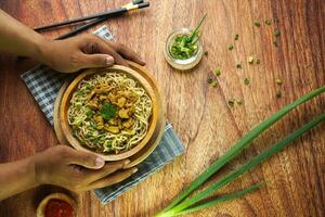 Nudeln mit Huhn, im Indonesien es ist bekannt wie mie ayam. serviert mit Grün Gemüse Chili Soße im ein Schüssel auf hölzern Hintergrund durch Hand foto