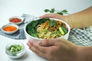 Nudeln mit Huhn, im Indonesien es ist bekannt wie mie ayam. serviert mit Grün Gemüse Chili Soße im ein Schüssel auf Weiß hölzern Hintergrund durch Hand foto