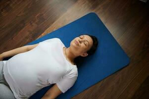 oben Aussicht von ein herrlich schwanger Frau Lügen auf ein Blau Yoga Matte während vorgeburtlich Entspannung Übungen beim Zuhause foto