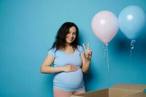 schwanger Frau lächelt Schlaganfälle Bauch zeigt an zwei Finger beim Kamera, Gefühl positiv Emotionen erwarten Zwillinge. Geschlecht Party foto