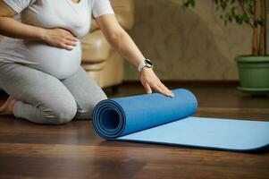 selektiv Fokus auf ein Blau Übung Matte auf ein hölzern Boden, und ein schwanger Frau bereit zum Schwangerschaft Yoga trainieren drinnen foto