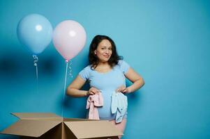 glücklich schwanger Frau halten Neugeborene Kleider auf ihr Bauch, lächelnd posieren mit Rosa und Blau Luftballons auf Blau Hintergrund foto