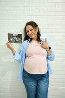 schwanger Frau lächelt beim das Kamera und Punkte gegenüber das Ultraschall beim ihr Baby, isoliert gegen ein Weiß Backstein Mauer. foto