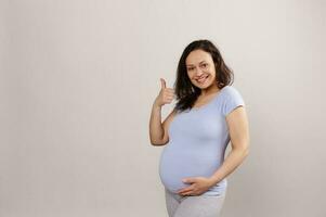 schön schwanger Frau im Blau T-Shirt sanft berühren streicheln ihr groß Bauch und zeigen Daumen oben suchen beim Kamera foto