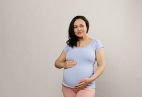 Porträt von schön Erwachsene schwanger Frau, gravid erwartungsvoll Mutter halten Hände auf ihr groß Bauch im spät Schwangerschaft foto