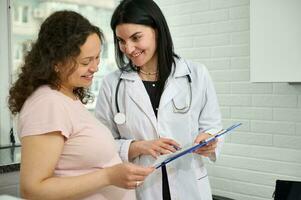 zuversichtlich weiblich Arzt tragen Weiß medizinisch Kleid, halten Zwischenablage, Beratung ihr Neu schwanger geduldig im Klinik foto