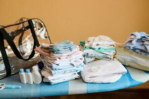 leeren Tasche in der Nähe ein Stapel von gebügelt Baby Kleidung, organisch Kosmetika auf Bügeln Planke, bereit zum Mutterschaft Krankenhaus foto