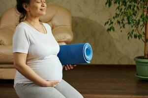glücklich Erwachsene schwanger Frau Sitzung im Held Pose, berühren ihr Bauch, genießen aktiv Fitness Lebensstil während Schwangerschaft foto
