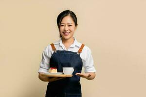 jung asiatisch Kellnerin Barista tragen Schürze Hand halten Kuchen und heiß Kaffee auf hölzern Tablett foto