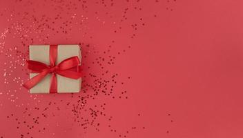 Geschenkbox eingewickelt in ein Bastelpapier mit rotem Band mit Schleife und Konfetti auf rotem Hintergrund monochrome festliche Wohnung lag mit Kopierraum