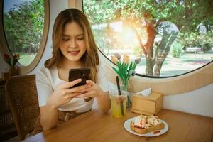 jung Erwachsene glücklich asiatisch Frau mit Clever Telefon zum Sozial Medien Anwendung, Einkaufen online, Transfer Geld, finanziell, Botschaft im Cafe. Hintergrund mit Fenster und warm Sonnenlicht auf Winter Jahreszeit. foto