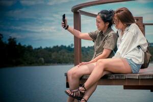 zwei froh heiter Mädchen nehmen ein Selfie während Sitzung zusammen auf das Brücke und suchen beim das schön Flüsse und Berge. zwei jung Asien Damen Rucksack zu Reise das Feiertage. entspannt. foto