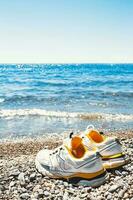 ein Paar von Weiß Turnschuhe auf das Kieselsteine von das Meer Ufer - - Laufen oder Gehen entlang das Ozean foto