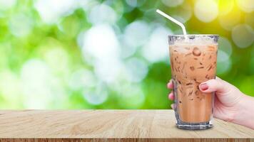 Hand halten das Glas vereist Kaffee auf hölzern Tabelle auf abstrakt verschwommen Grün Bokeh Hintergrund, Eis Latté Kaffee foto