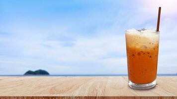 vereist thailändisch Milch Tee im Glas, Milch Eis Tee, Cheddar ist ein traditionell thailändisch trinken auf hölzern Tabelle mit Strand Landschaft Natur Hintergrund, Sommer- Getränke mit vereist foto