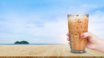 Hand halten das Glas vereist Kaffee auf hölzern Tabelle mit Strand Landschaft Aussicht Natur Hintergrund, Eis Latté Kaffee foto